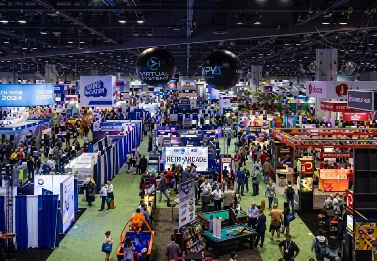 Large tech trade show floor with gaming booths, startup exhibits, hanging signage, and attendees exploring convention center