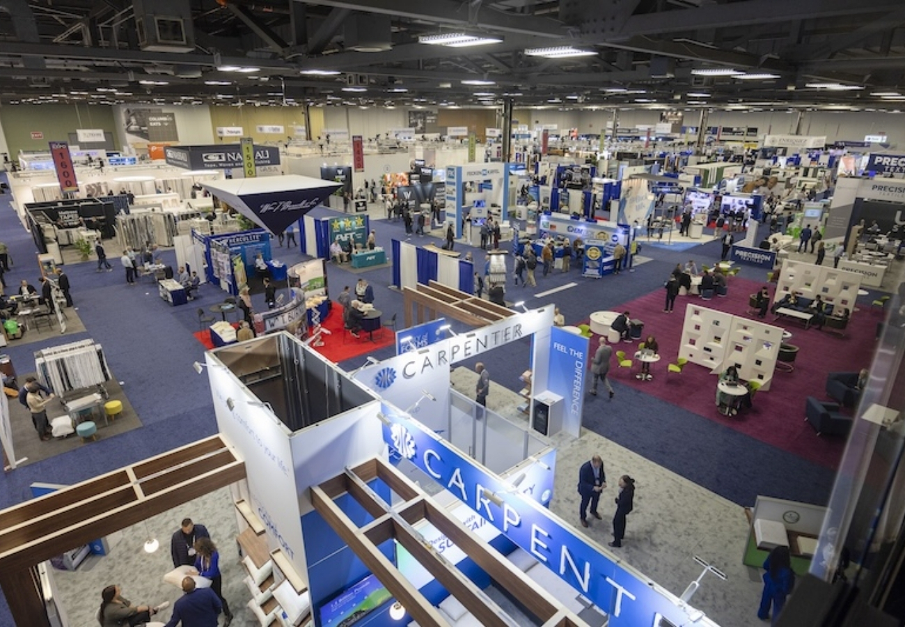 Large convention center trade show floor with multiple exhibit booths, branded displays, seating areas, and attendees walking through aisles