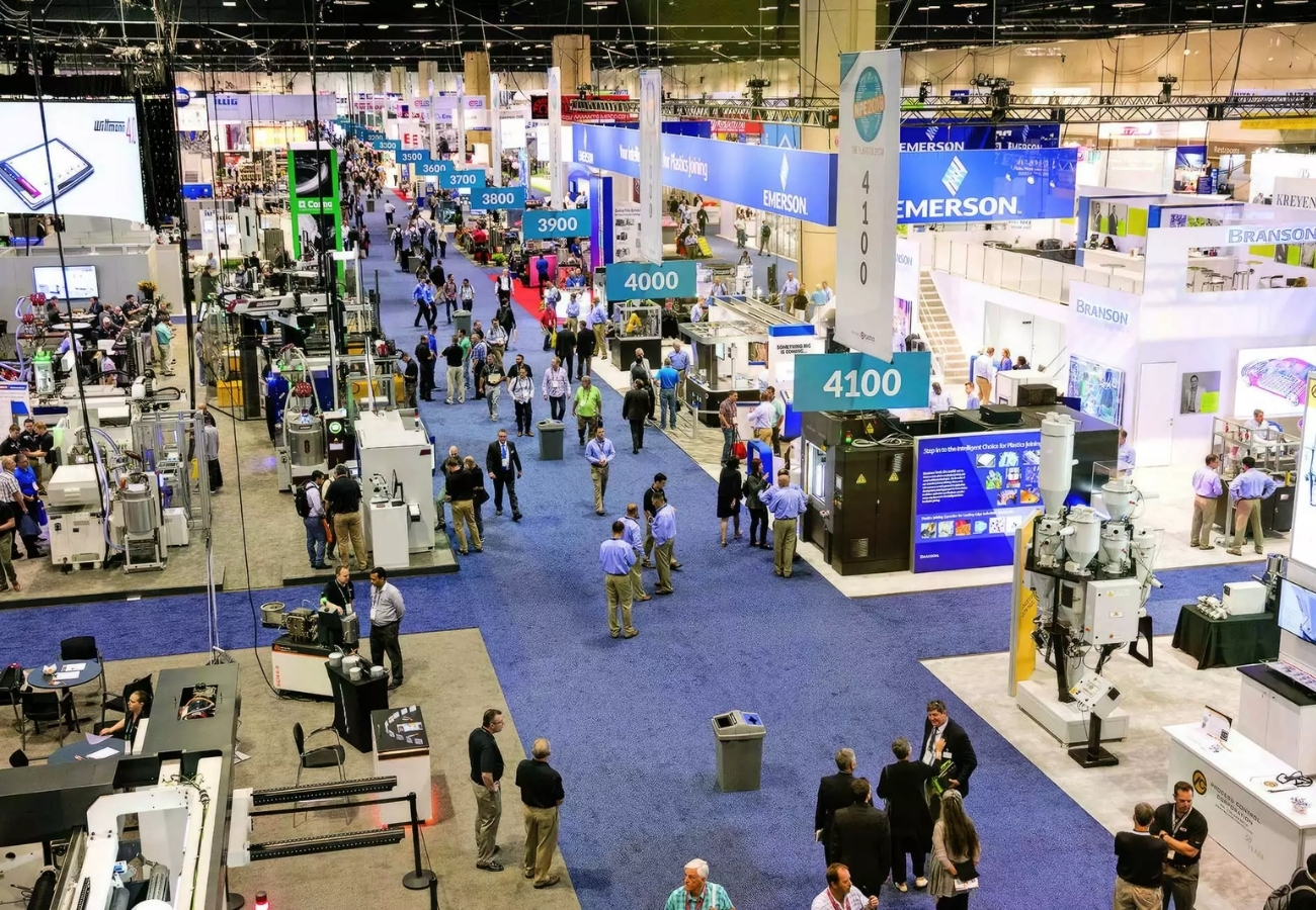 Busy trade show floor with multiple exhibit booths, numbered aisles, industrial displays, and attendees networking at large convention center